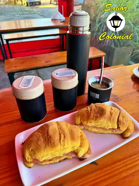 Equipo matero con termo de acero inoxidable, yerbera, azucarera, mate con bombilla y medialunas rellenas en mesa con bancos – Sabor Colonial